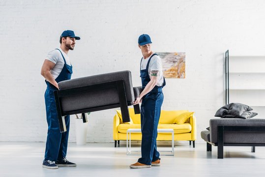 Two professional movers in blue uniforms carrying a black armchair inside a modern living room, showcasing efficient Furniture Removal services kent wa during a residential move.